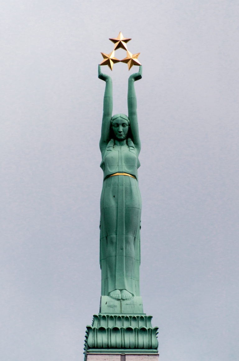 Green-patinated Liberty figure atop Riga’s Freedom Monument holding three gilded stars against the sky.