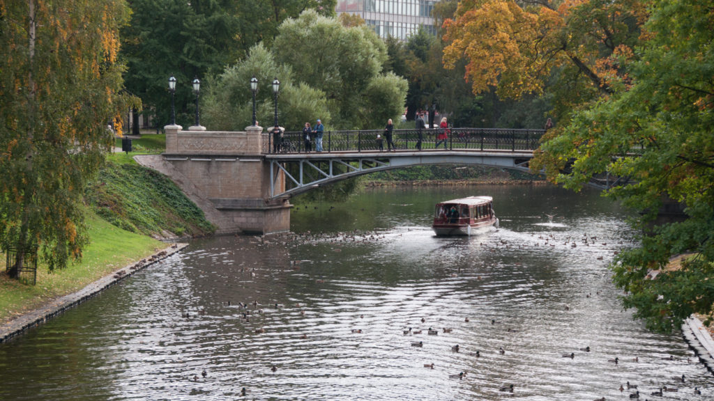 Small tour boat on the Riga City Canal approaching a low bridge among trees and autumn foliage.