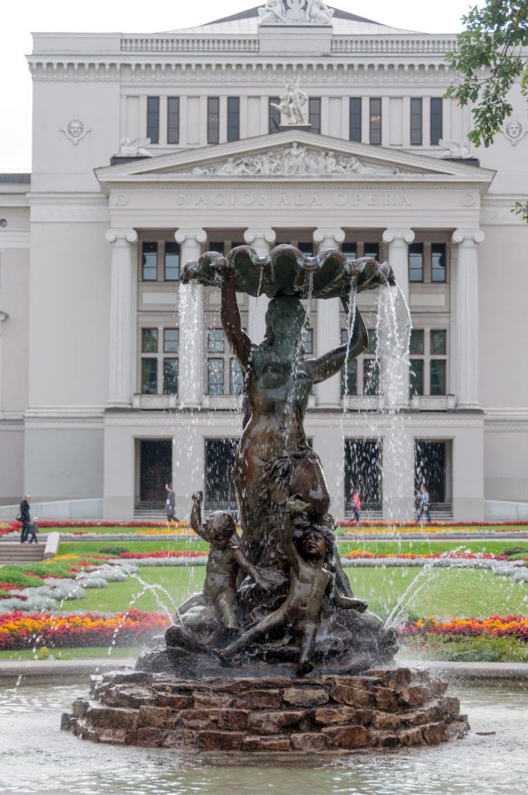 Close frontal view of the bronze Nymph Fountain with water jets and the Latvian National Opera behind it.