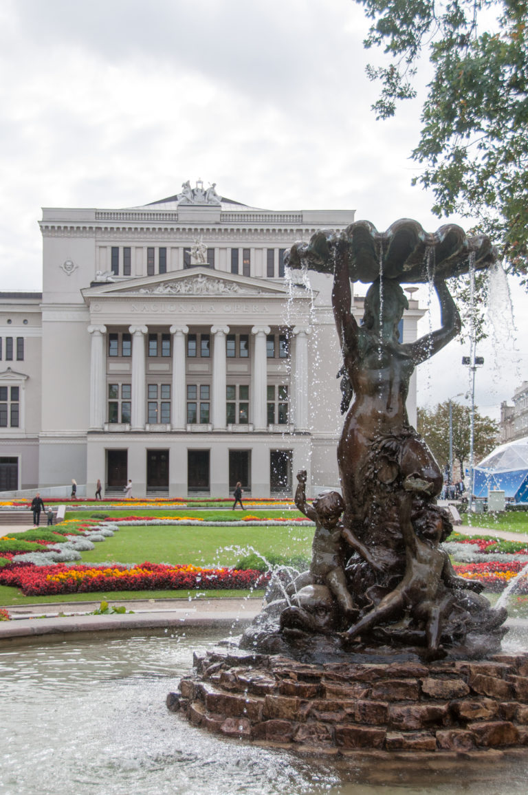 Nymph Fountain spraying water in front of colorful gardens and the Latvian National Opera facade in Riga.