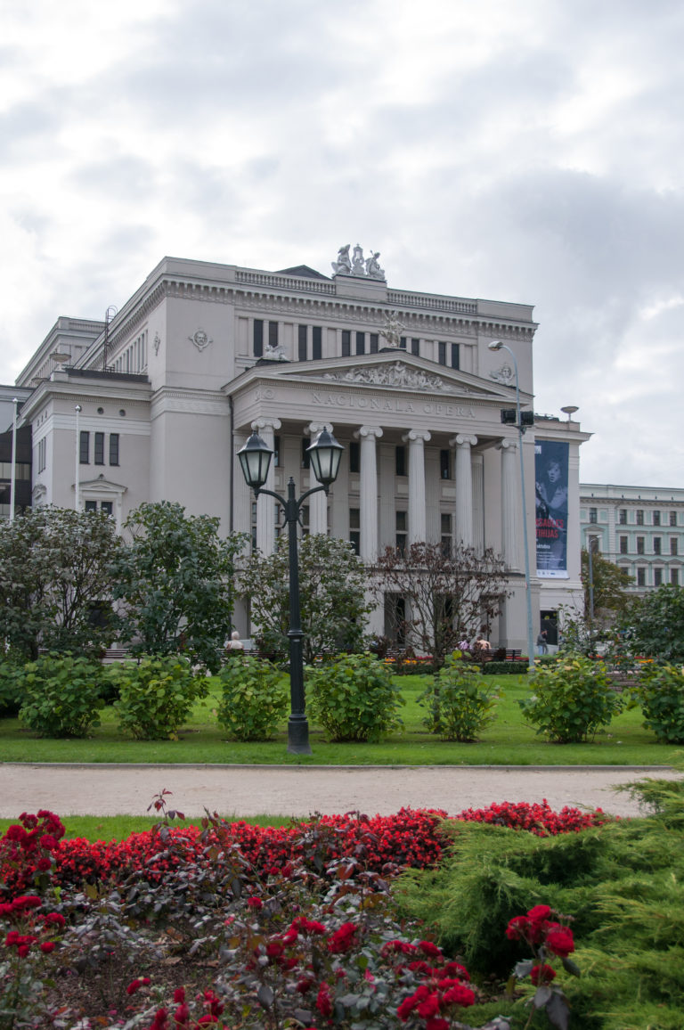 Latvian National Opera seen across formal gardens with flower beds, lawns, lamps, and a columned classical facade.