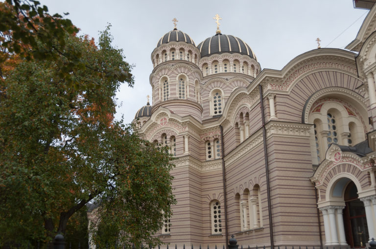 Nativity of Christ Cathedral in Riga with striped masonry, arched windows, domes, and a leafy tree in front.