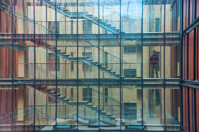 Glass-walled modern atrium with stacked staircases, reflections, metal framing, and a person on an upper level.