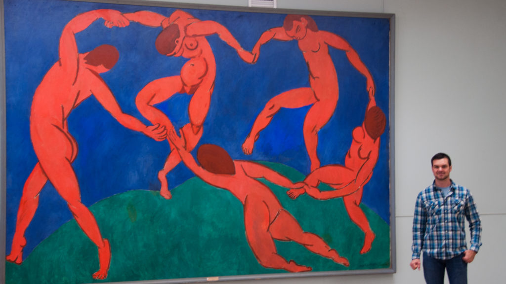 Museum visitor beside Henri Matisse’s monumental Dance, with five red figures circling across green ground beneath deep blue.