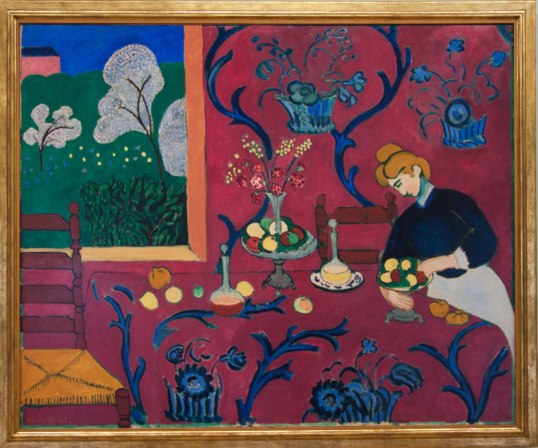 Woman arranging fruit at a table in Matisse’s red interior covered with blue floral arabesques.