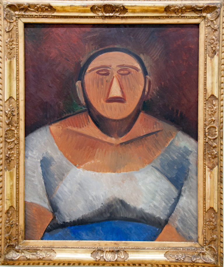 Pablo Picasso’s 1908 Farm Woman, a frontal bust with a mask-like face, triangular nose, broad brown neckline, and pale blue-gray blouse.