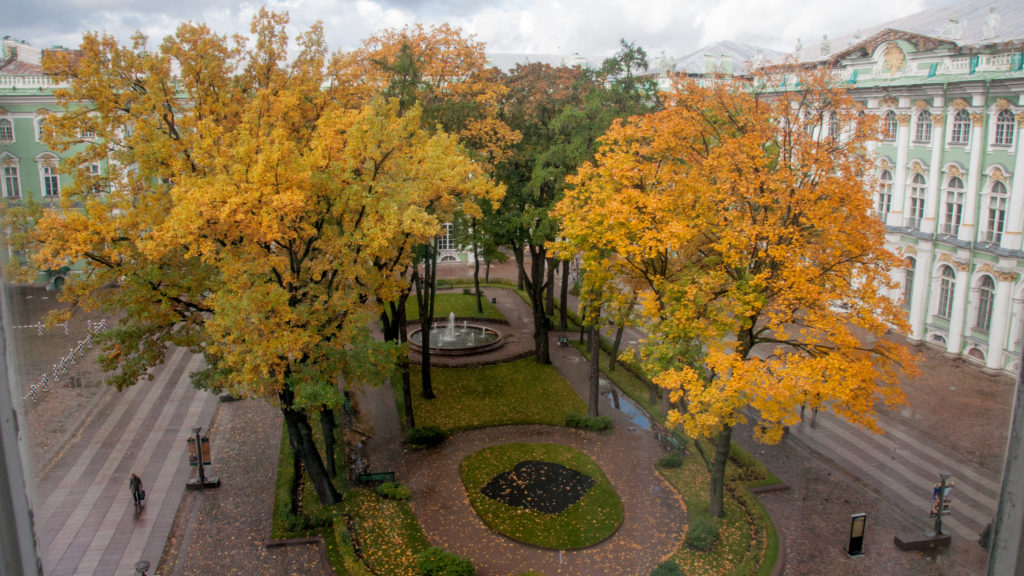 Elevated view of an autumn courtyard garden with golden trees, curving paths, fountain, and green-and-white palace façades.