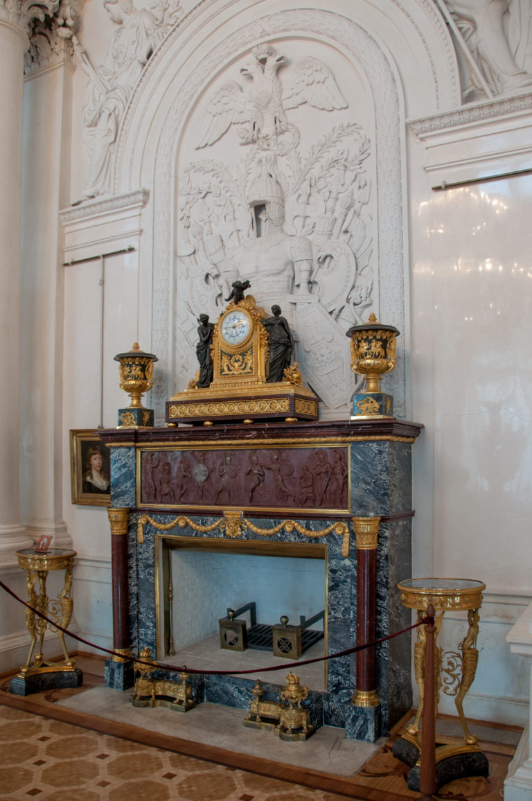 Gilt mantel clock and matching decorative vessels on a marble console before a large white relief panel.