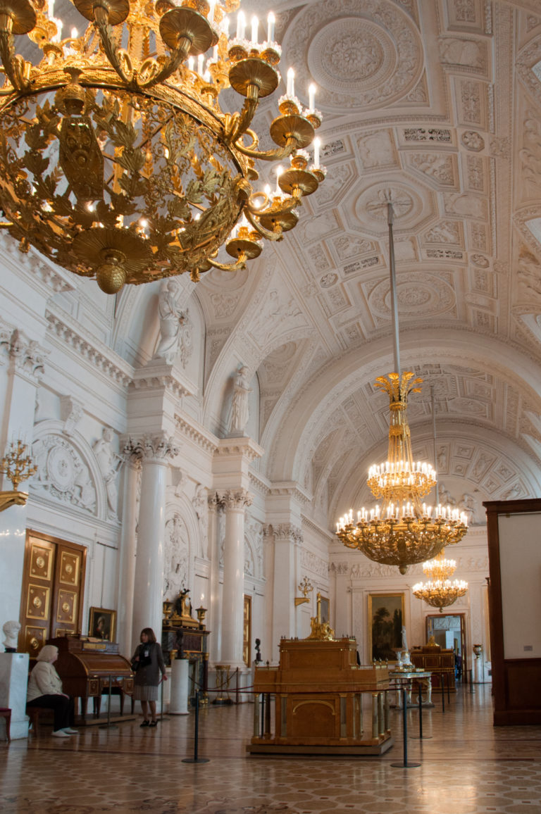 Grand white palace gallery with gilded chandeliers, columns, ornate ceiling, wood floor, and museum visitors.