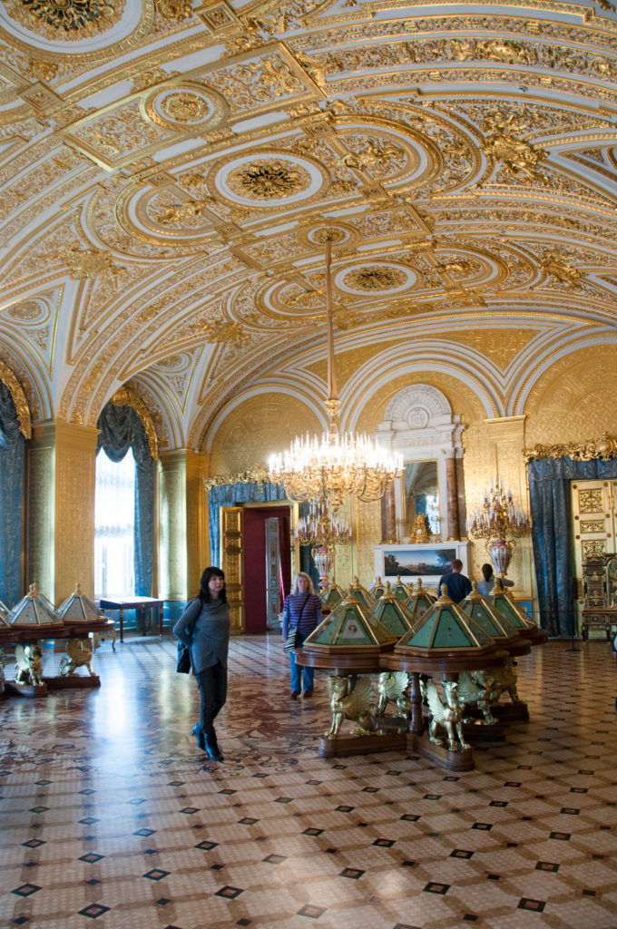 Ornate Hermitage gallery with gilded vaulted ceiling, chandeliers, parquet floor, visitors, and rows of glass display cases.