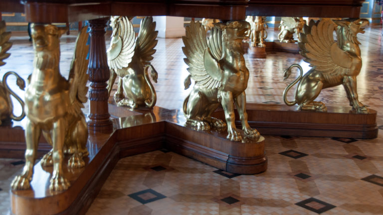 Gilded winged-creature supports beneath a dark wooden display table, reflected in polished surfaces above a patterned floor.