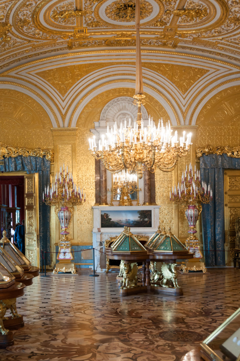 Gilded Hermitage gallery centered on a white marble fireplace and chandelier, with decorative vessels and glass display cases.