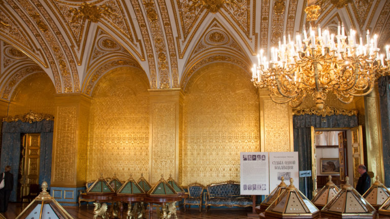 Gilded vaulted Hermitage room with large chandelier, blue seating, wall ornament, and small display cases.