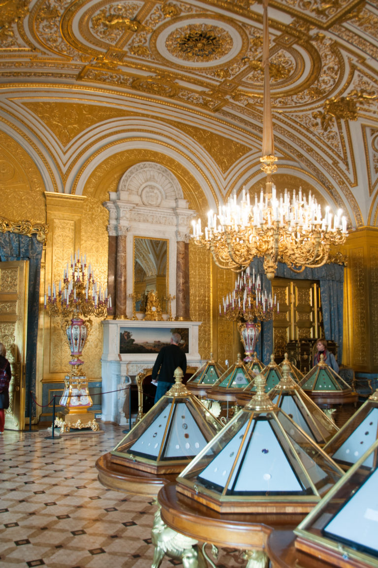 Rows of low glass display cases lead toward a marble fireplace and chandelier in an ornate gilded Hermitage gallery.