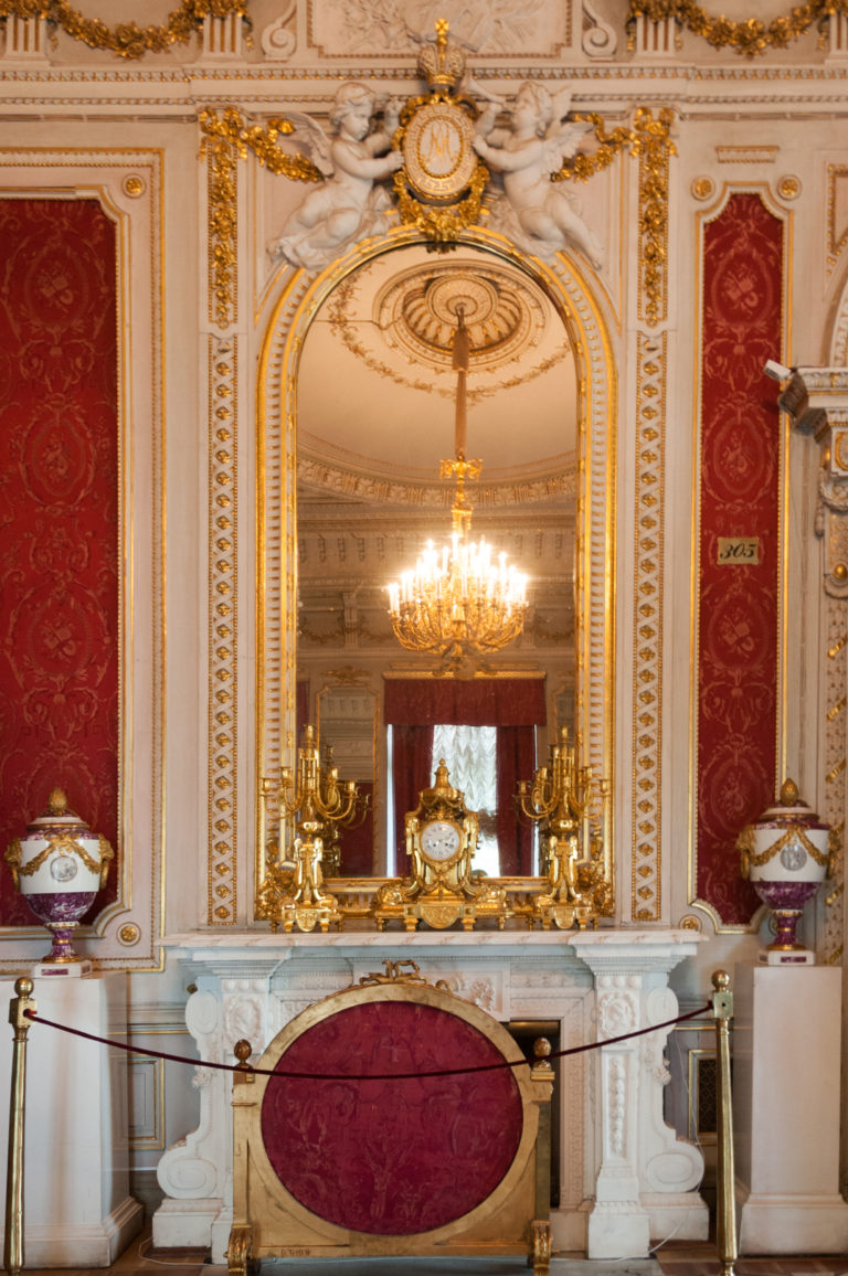 Red-and-white Hermitage interior with marble mantel, central clock, gilded ornaments, arched recess, and chandelier.