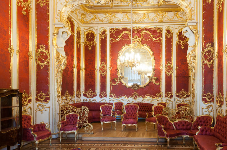 Red-and-gold Hermitage salon with oval mirror, gilded wall ornament, chandelier, and matching upholstered chairs and sofas.