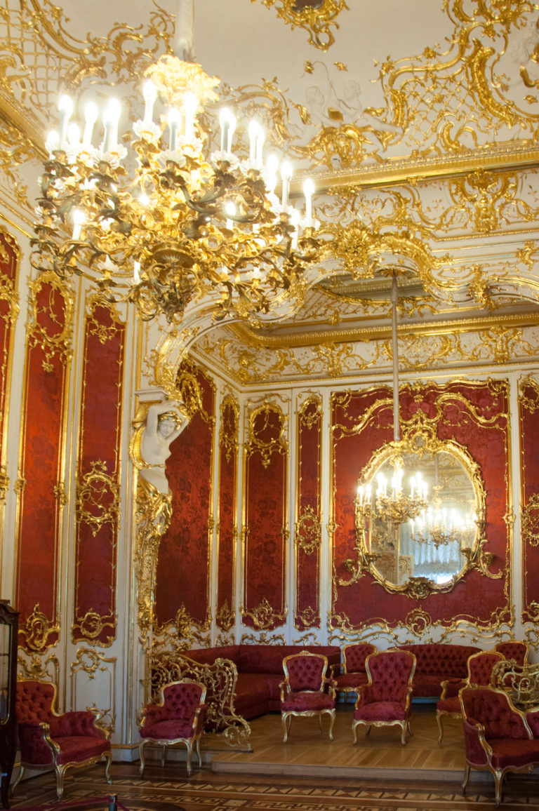 Wide view of a red-and-gold Hermitage salon with two chandeliers, oval mirror, gilded panels, and red upholstered seating.