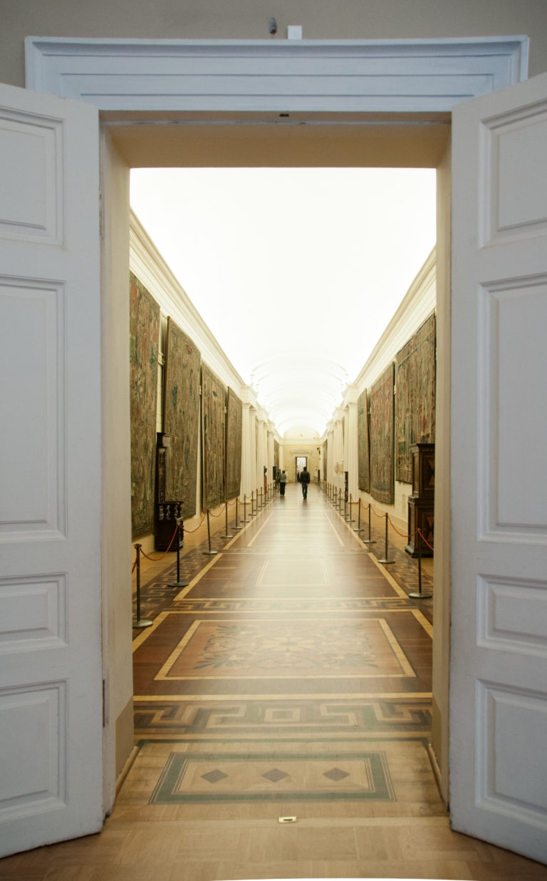 Long Hermitage corridor seen through open doors, with large tapestries lining both walls and visitors in the distance.