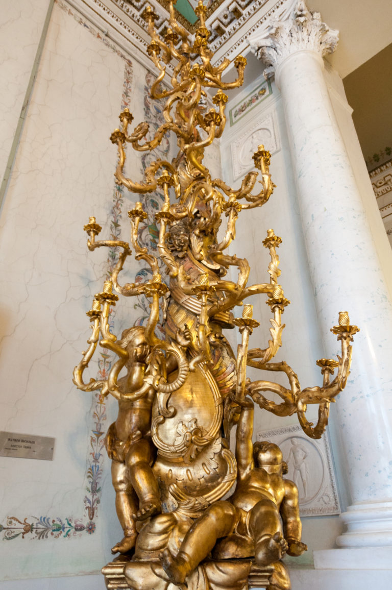 Large gilded clock-and-candelabrum form with scrolling branches and figures beside a pale marble column.