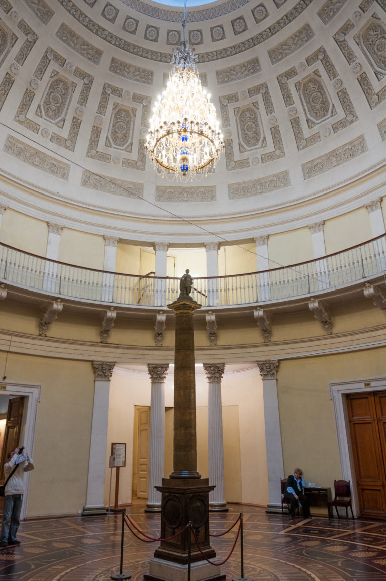 Circular Hermitage hall with upper gallery, crystal chandelier, and tall dark column monument centered on the floor.