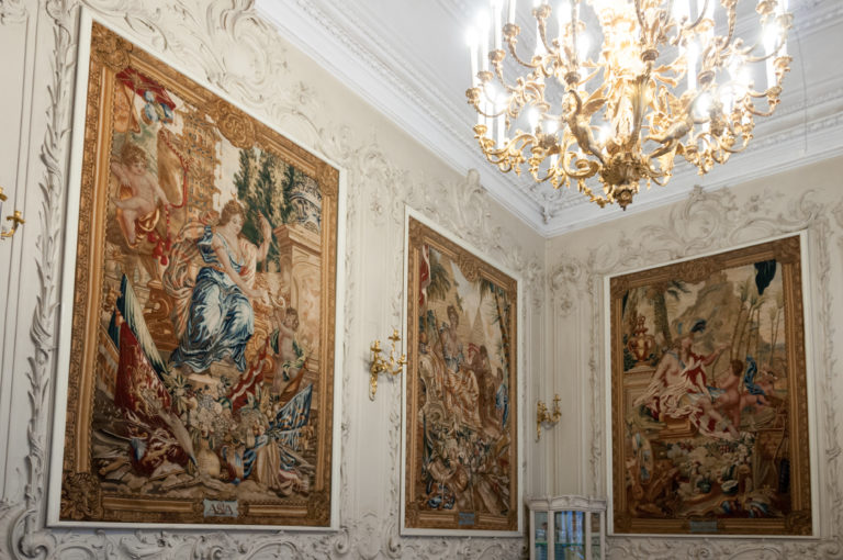 Bright Hermitage room with three large narrative tapestries, white relief wall decoration, and a gilded chandelier.