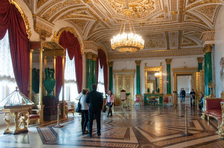 Wide Malachite Room in the Winter Palace with green malachite columns, red draperies, gilded ceiling ornament, monumental vases, and patterned parquet.
