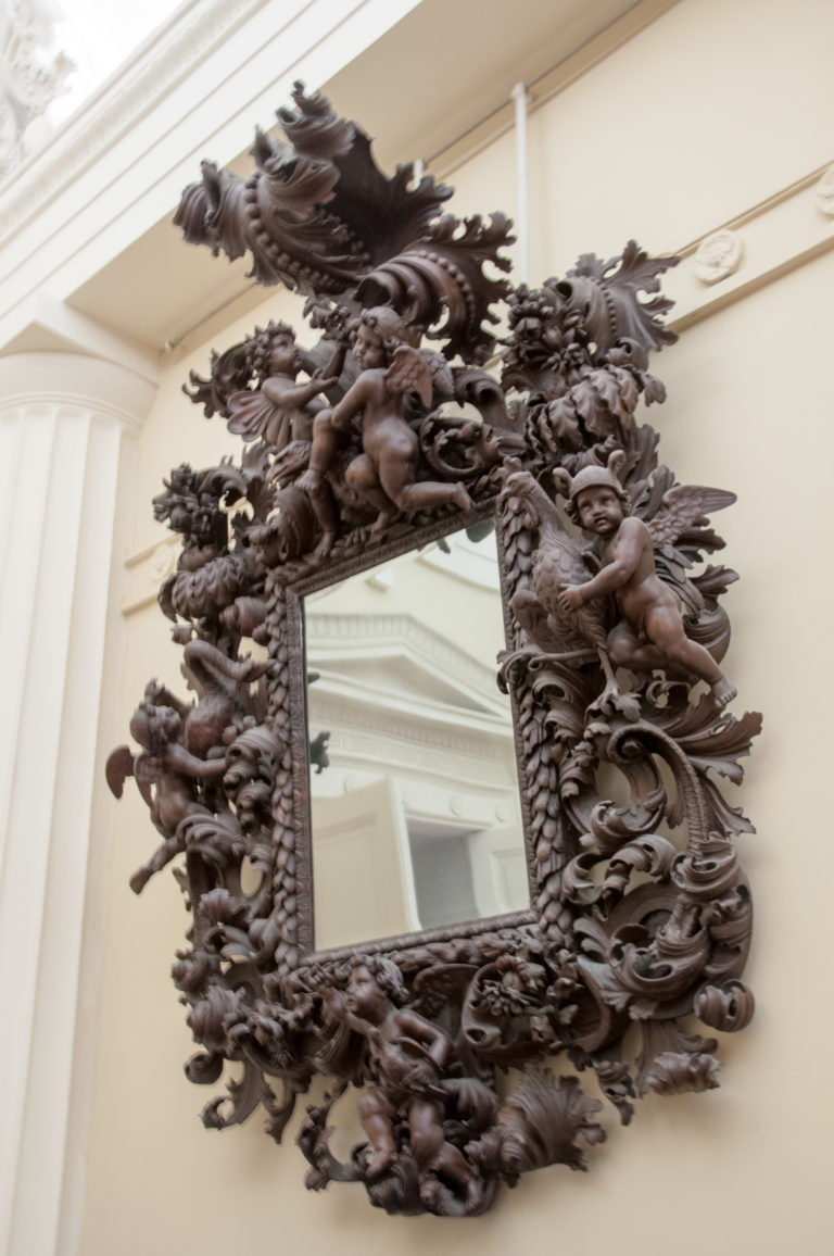 Rectangular mirror in a dense dark carved-wood frame of scrolling foliage and sculptural ornament against a pale wall.