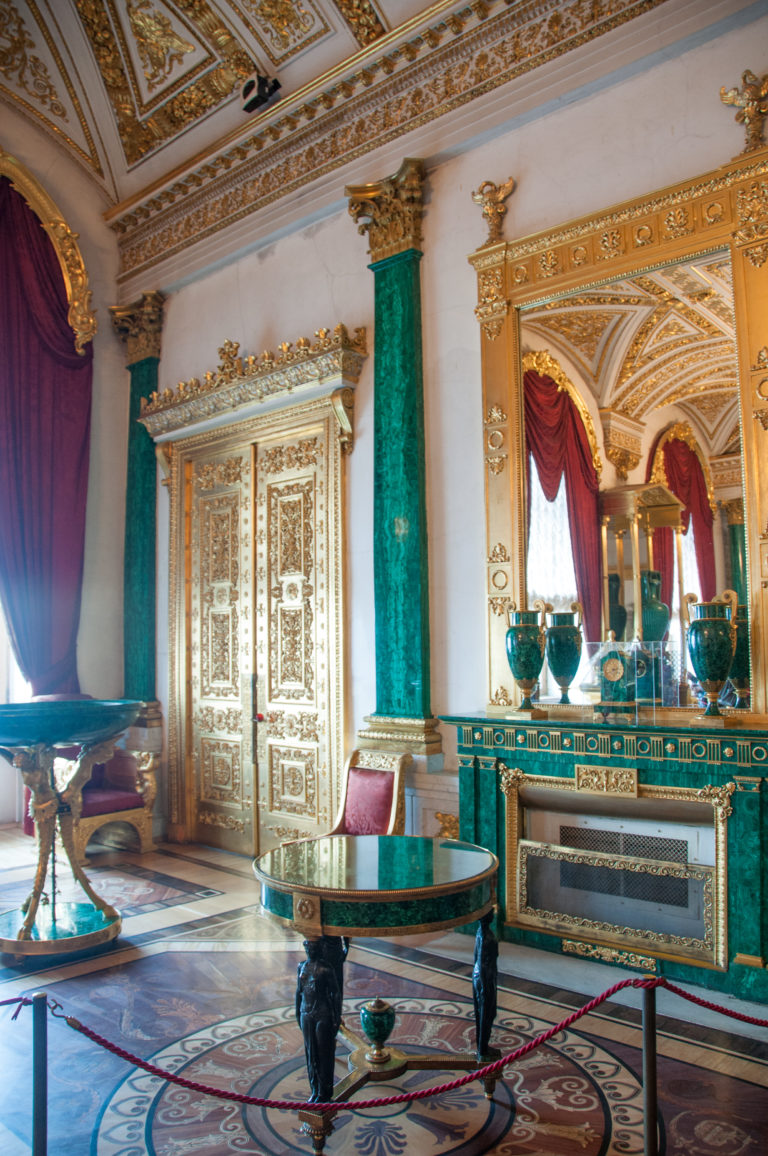 Malachite Room detail with green stone fireplace and columns, gilded mirror and doors, circular table, and reflected green vases.