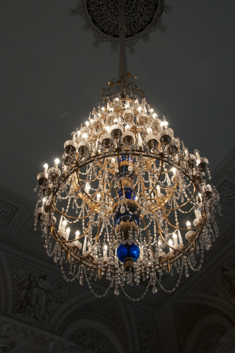Large illuminated chandelier viewed from below, with radiating branches, crystal drops, and distinctive blue glass accents.