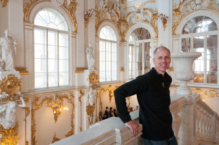 Visitor on the Jordan Staircase landing amid tall arched windows, white sculpture, mirrors, gilded ornament, and red carpet.