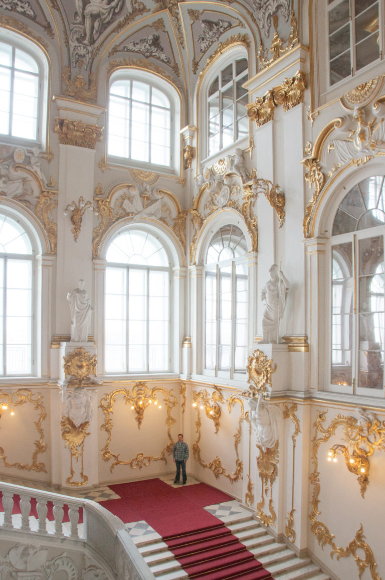 Jordan Staircase in the Winter Palace with arched windows, mirrors, allegorical statues, white balustrades, gilded ornament, and red carpet.