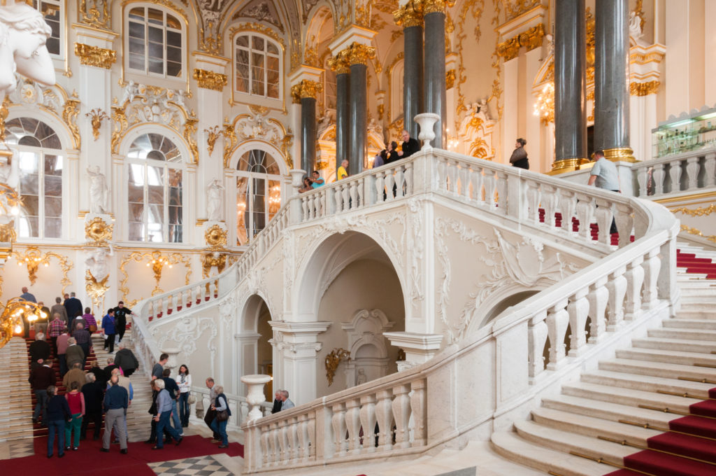 Broad Jordan Staircase view with branching white marble flights, dark columns, gilded capitals, red carpet, and groups of visitors.
