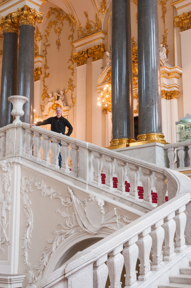 Close Jordan Staircase detail with curving white balustrade, carved relief, dark columns, gilded capitals, and a visitor beyond.