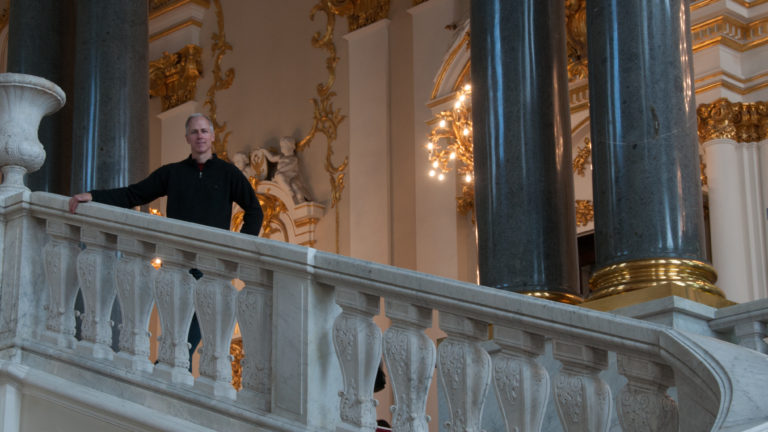 Visitor framed by dark columns behind the white balustrade of the Jordan Staircase, with gilded decoration and chandelier light beyond.