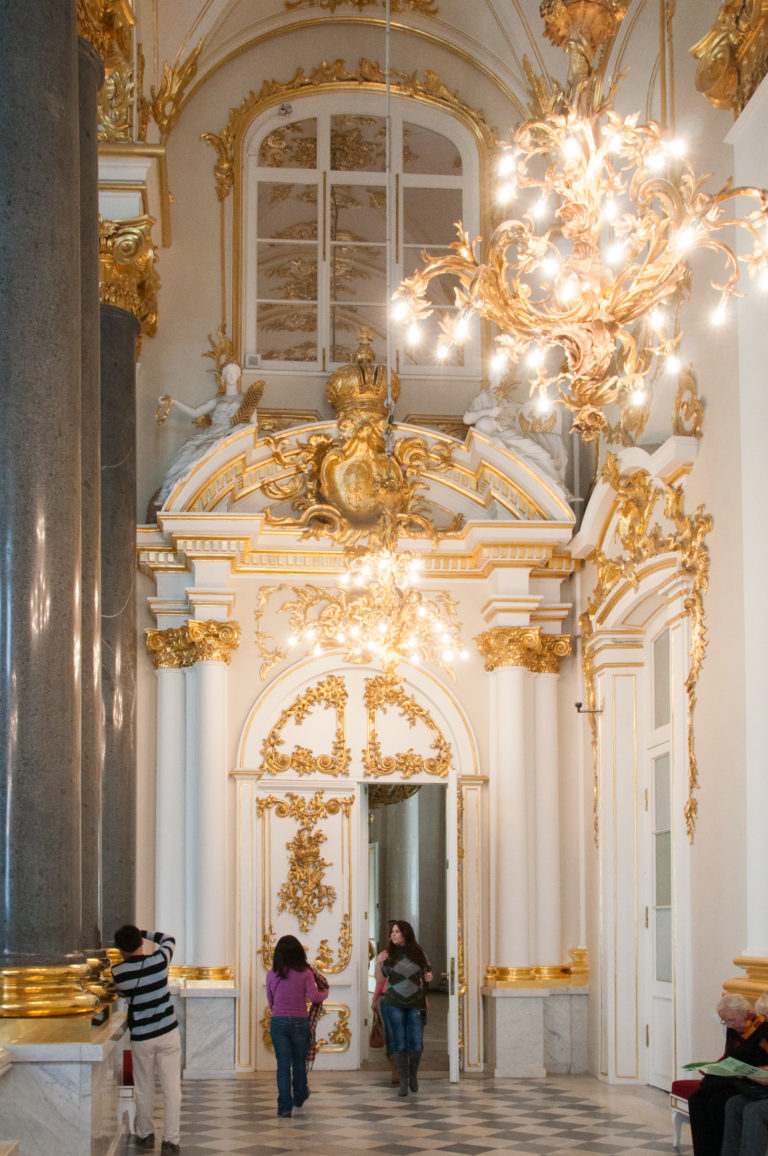 White-and-gold Hermitage doorway beneath a large chandelier, with dark columns, gilded carving, and several visitors below.