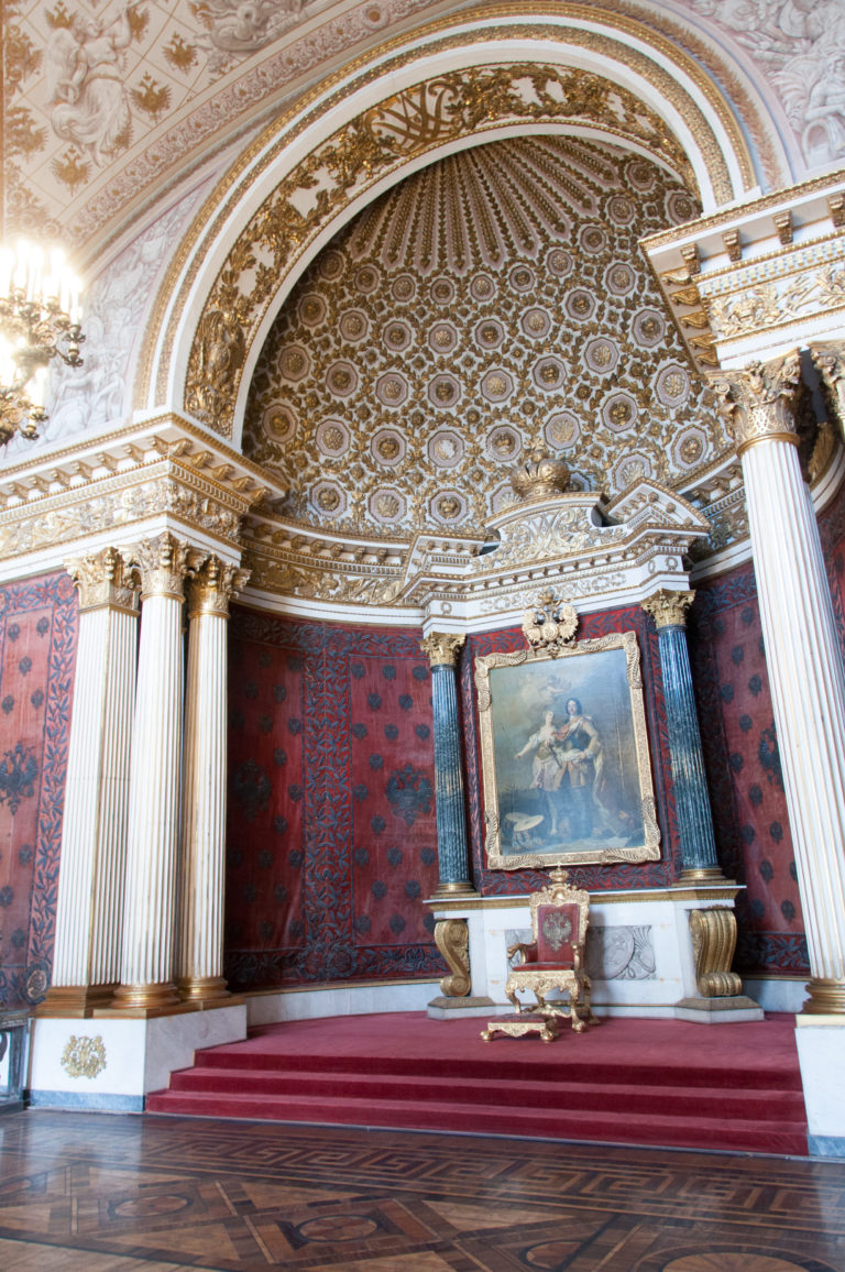 Red ceremonial alcove with fluted white columns, patterned half-dome, central painting, gilded furnishings, and raised carpeted platform.