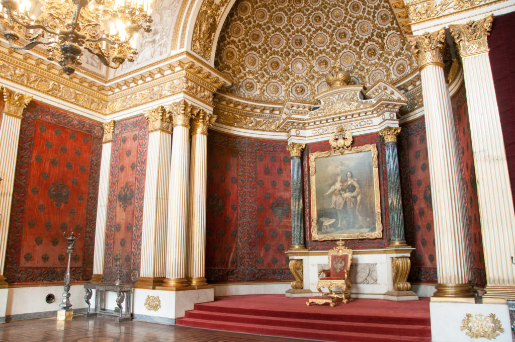 Closer angled view of a red ceremonial alcove with monumental painting, white columns, patterned vault, and gilded central furnishing.