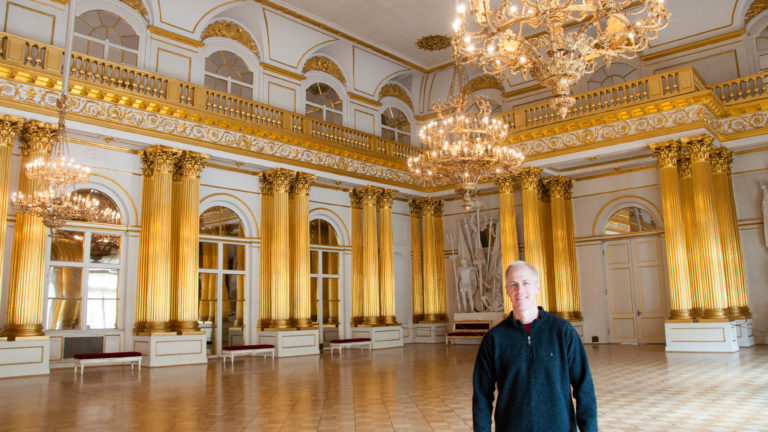 Visitor in the Armorial Hall surrounded by gilded fluted columns, chandeliers, arched openings, upper gallery, and parquet floor.