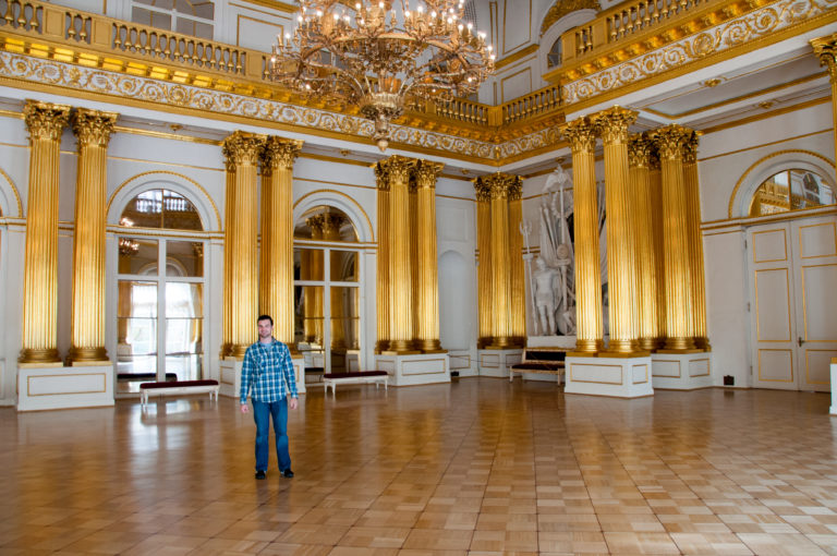 Solitary visitor in the Armorial Hall with towering gilded columns, chandeliers, arched openings, upper gallery, and parquet floor.