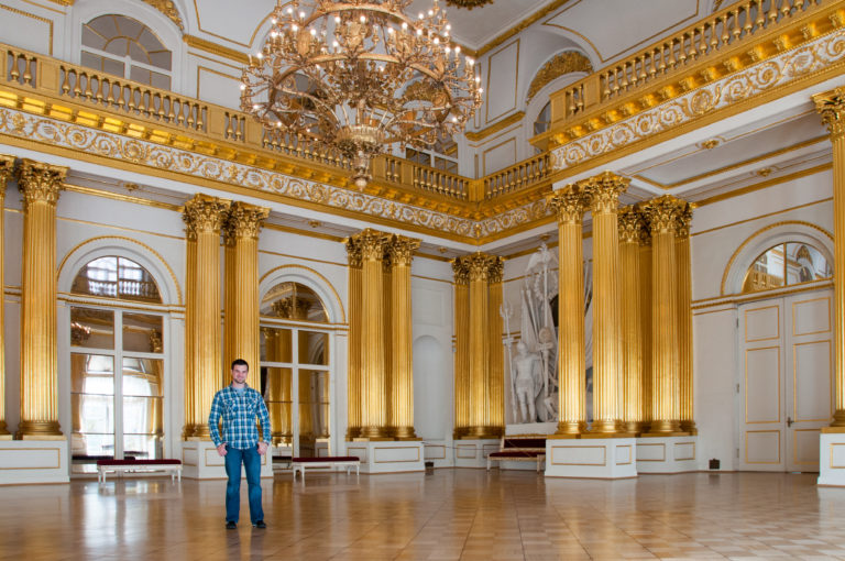 Visitor framed by gilded columns and chandeliers in the Armorial Hall, with arched openings and upper gallery behind.