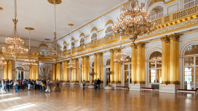 Visitors gathered in the Armorial Hall beneath chandeliers and gilded columns, with upper gallery and sunlit parquet floor.