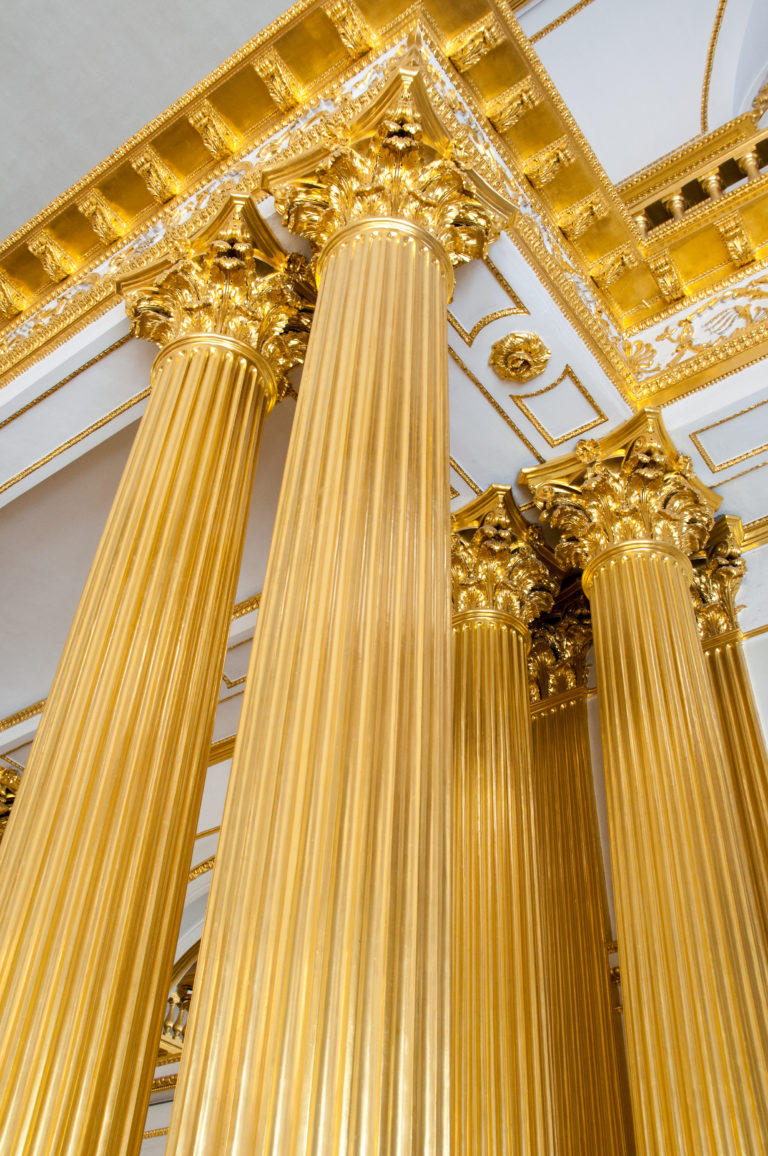 Low-angle view up gilded fluted columns to ornate leafy capitals and gold-trimmed entablature in the Armorial Hall.