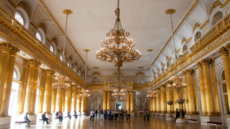 Long axial view of the Armorial Hall with repeated gilded columns, chandeliers, upper galleries, arched openings, and visitors.