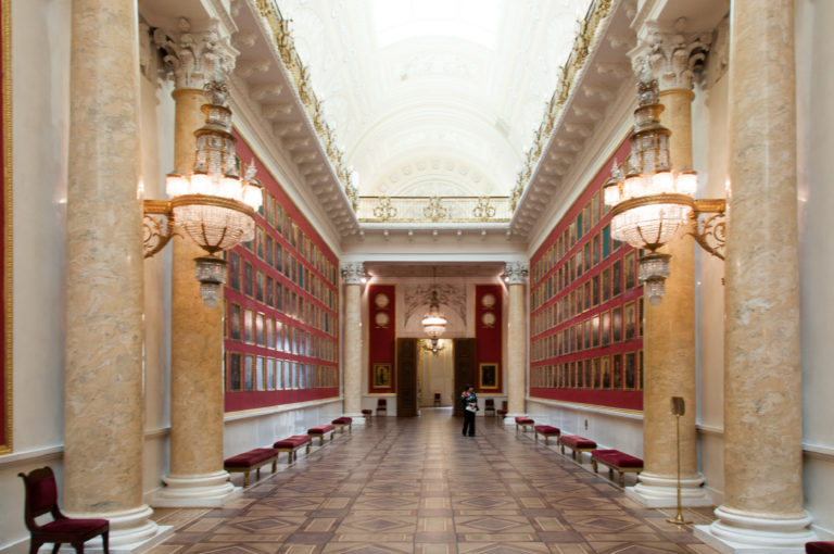 Long Hermitage gallery with red walls densely hung with small portraits, stone columns, hanging lamps, benches, and parquet floor.