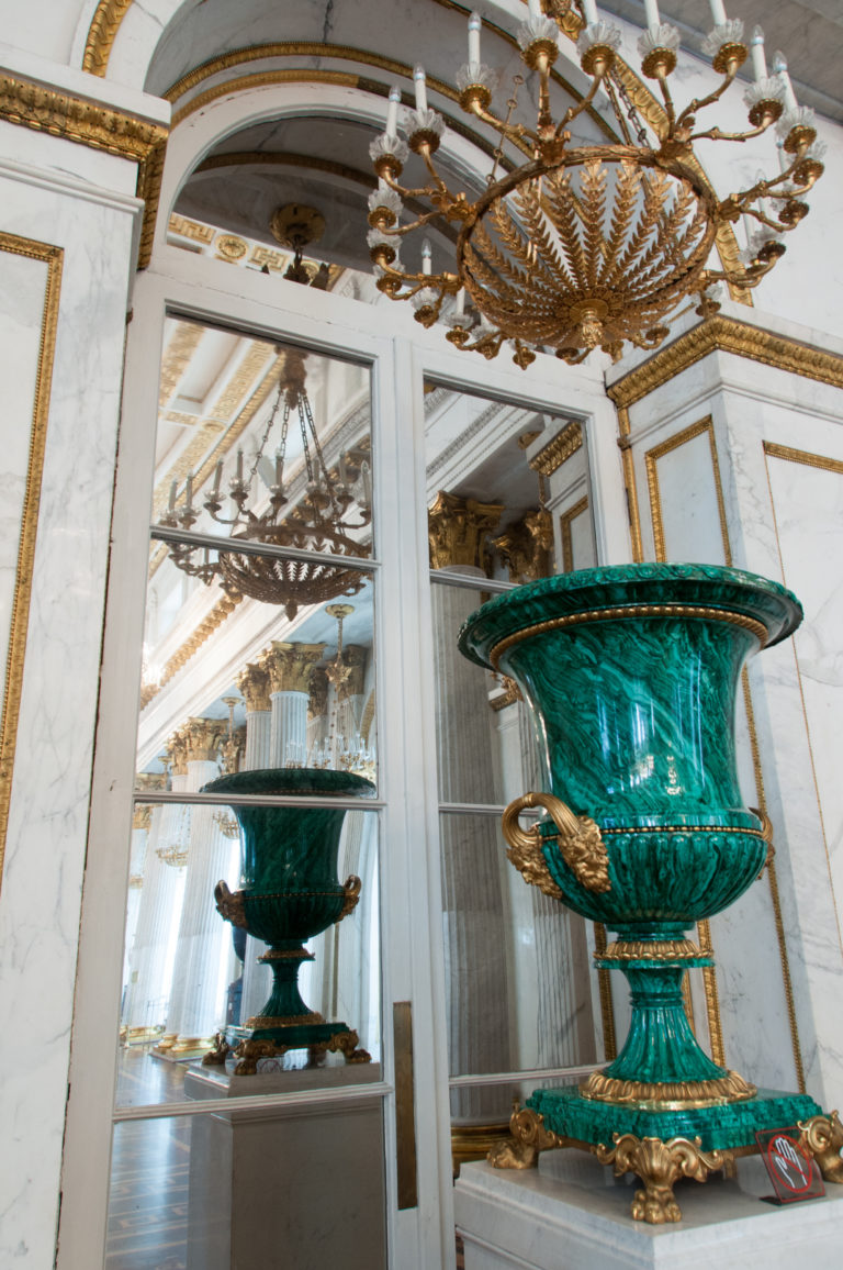 Monumental polished green vase with flared rim and gilded mounts reflected in mirrored wall panels beneath a chandelier.