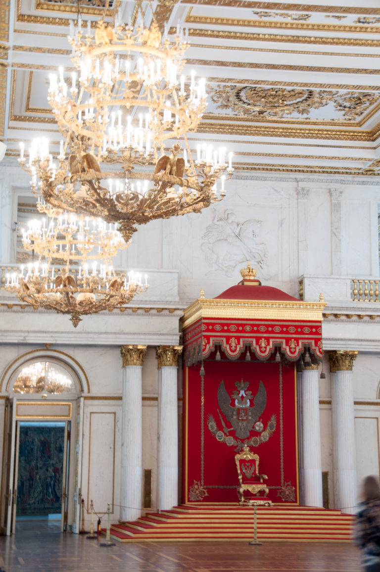 Raised red throne dais and canopy with double-headed eagle, framed by white columns and multiple chandeliers in the Hermitage.