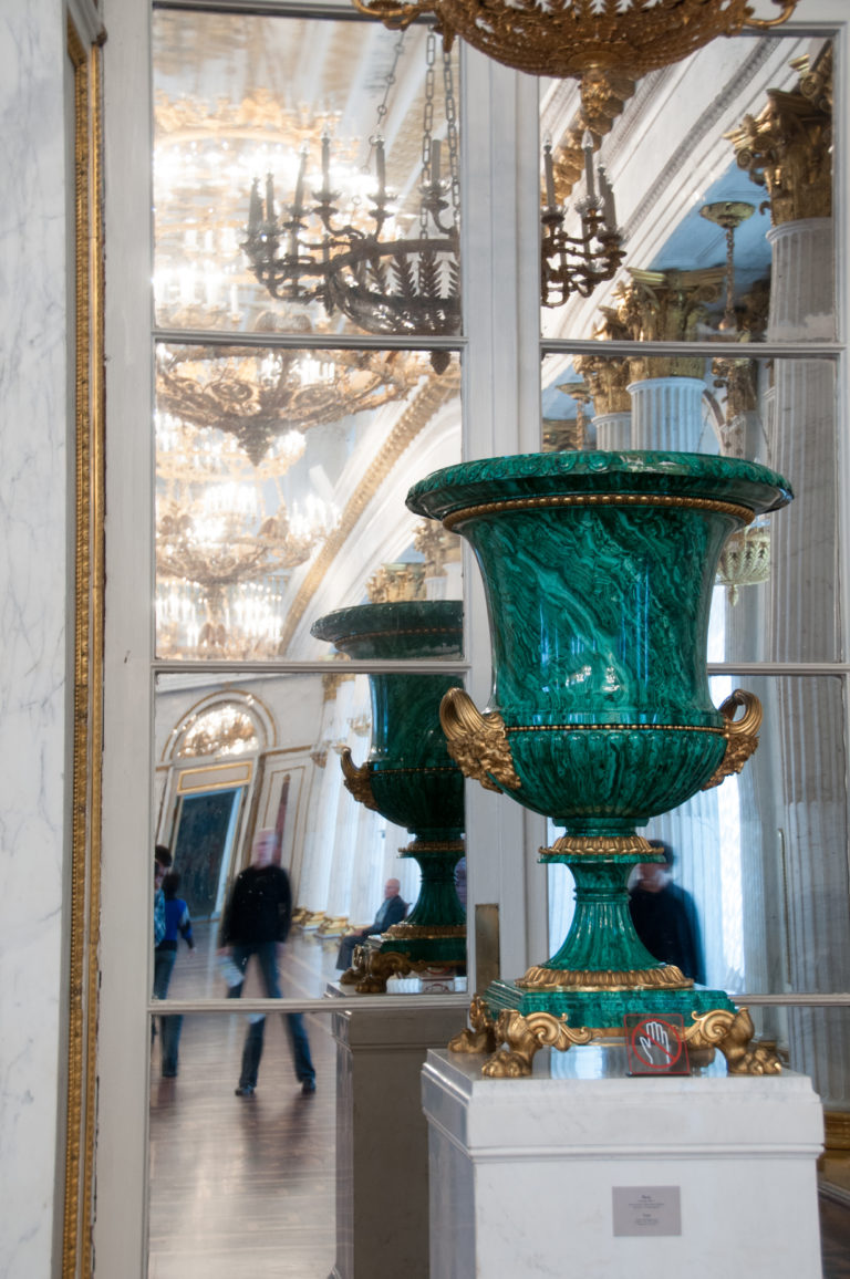 Front view of a large polished green vase with gilded handles and base, reflected in mirrors with visitors behind.