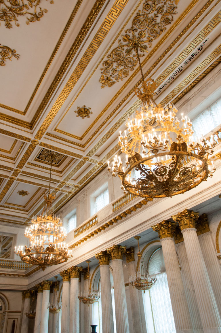 Multiple chandeliers hanging beneath an ornate white-and-gold ceiling above fluted columns and high windows in the Hermitage.