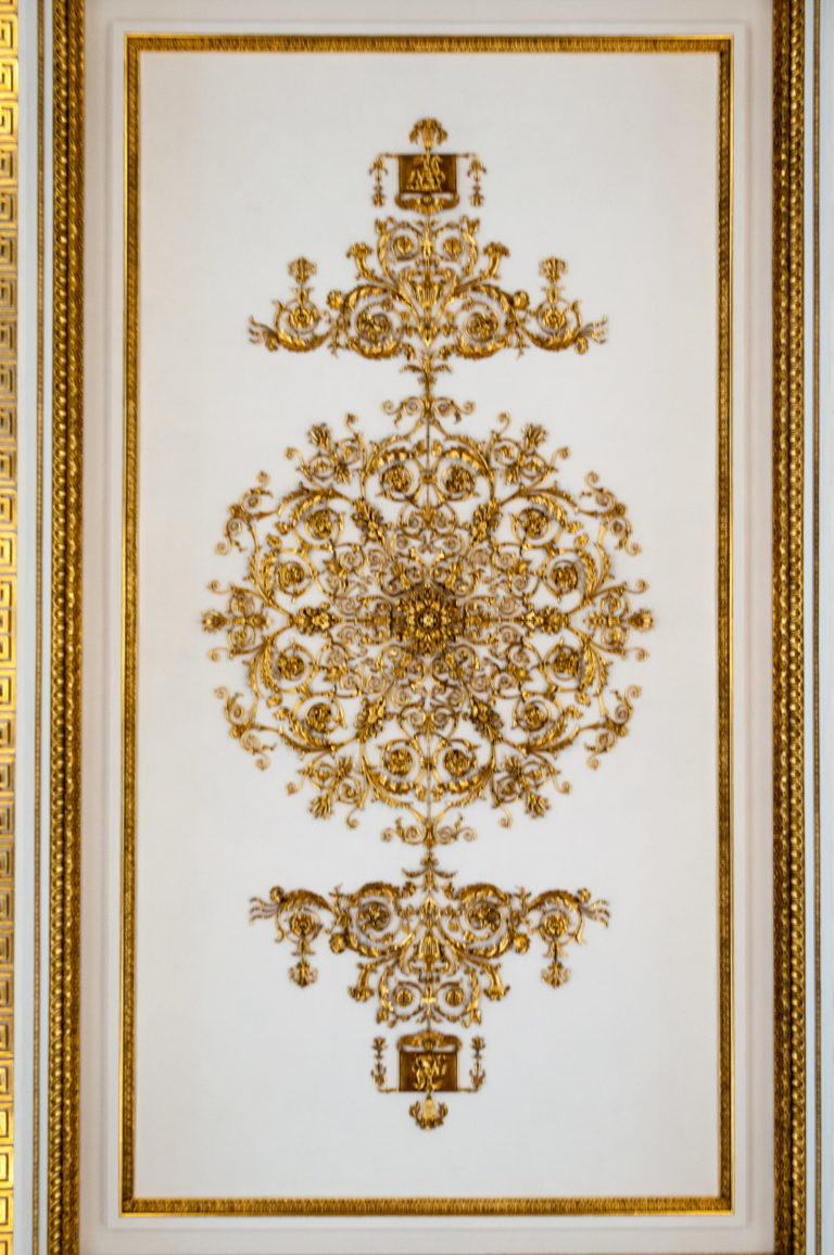 Symmetrical gilded arabesque of scrolling foliage and floral motifs centered within a rectangular gold border on a pale panel.