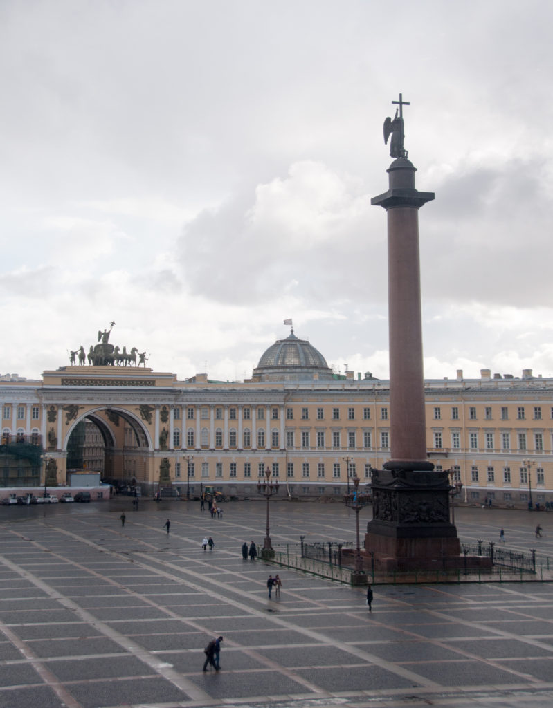 Alexander Column on Palace Square with the pale-yellow General Staff Building and triumphal arch behind under cloudy sky.
